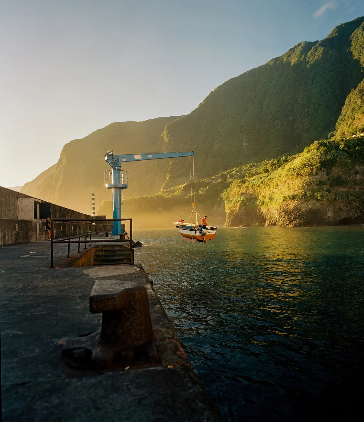 A small boat hangs from a crane beside a concrete quay, with sunlit cliffs beyond.