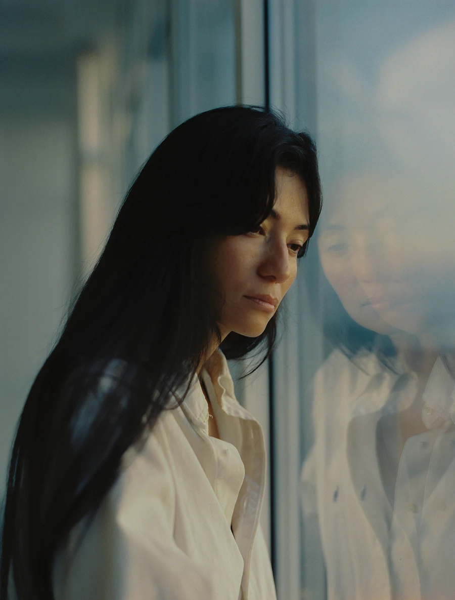 A person with long dark hair stands beside a window, reflected in the glass.